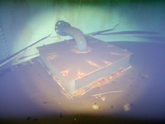 A mesh strainer box on the floor of a steel water tank, surrounded by settled sediment.